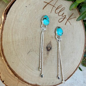 May include: A pair of silver dangle earrings featuring teardrop-shaped turquoise stones with dark matrix veining. Each earring has a polished silver setting and two long, slender silver wires that hang down from the stone, ending in small silver spheres.