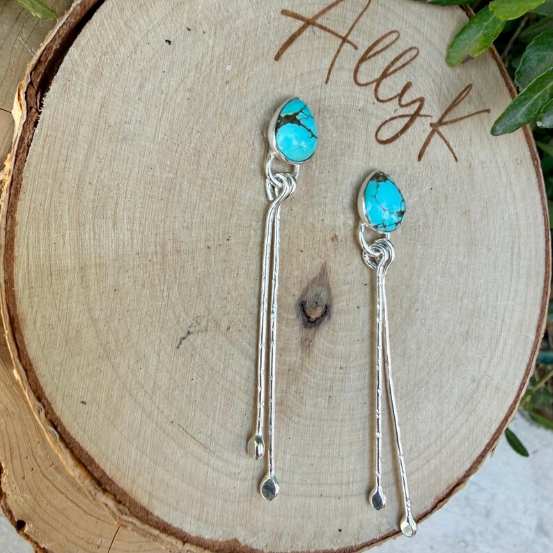 May include: A pair of silver dangle earrings featuring teardrop-shaped turquoise stones with dark matrix veining. Each earring has a polished silver setting and two long, slender silver wires that hang down from the stone, ending in small silver spheres.