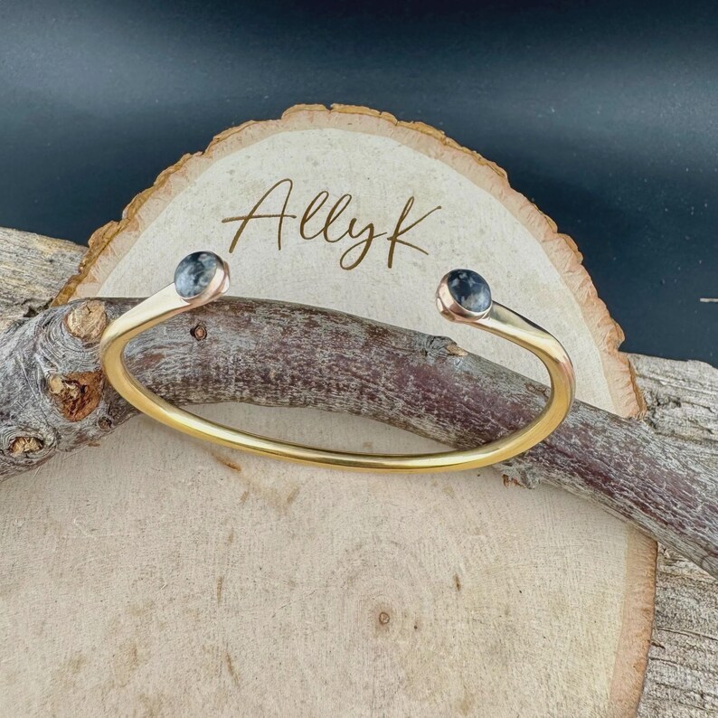 May include: A gold-colored cuff bracelet with a curved design. The bracelet features a small, round, dark stone at each end. The bracelet is displayed on a wooden surface with the name "Allyk" written on a wooden slice.