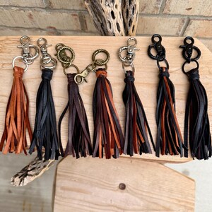 May include: A collection of leather tassel keychains in various colors, including brown, black, and tan. Each keychain features a metal clasp and a bundle of fringed leather strips. The keychains are displayed on a wooden surface.