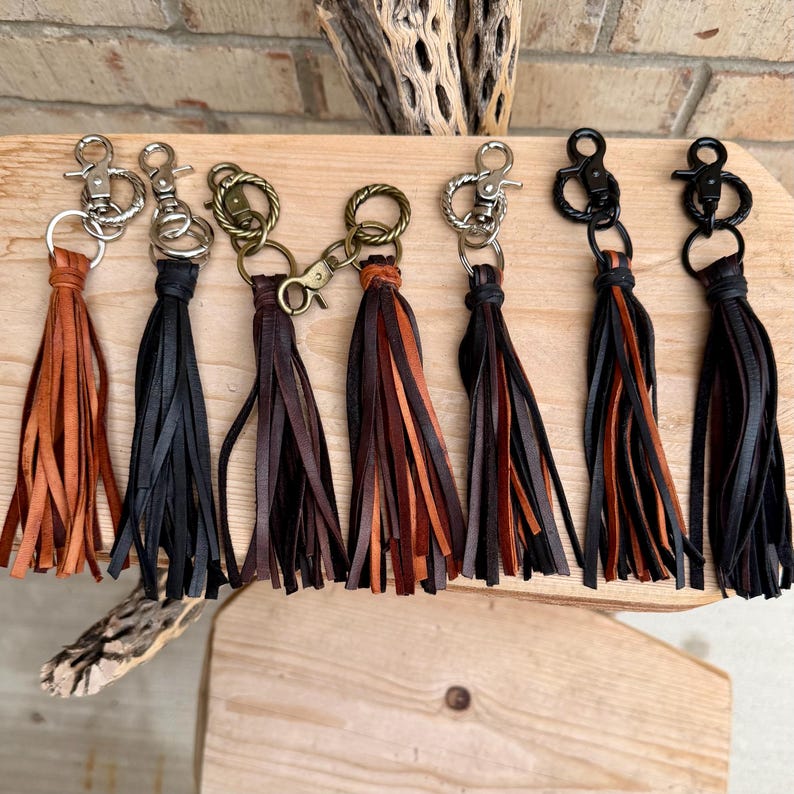 May include: A collection of leather tassel keychains in various colors, including brown, black, and tan. Each keychain features a metal clasp and a bundle of fringed leather strips. The keychains are displayed on a wooden surface.