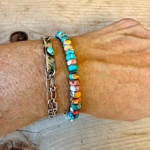 Pode incluir: Um conjunto de pulseiras composto por um fio de contas redondas multicoloridas em tons de turquesa, laranja e branco, e uma segunda pulseira com uma barra met&aacute;lica retangular com uma pedra turquesa incrustada numa corrente de elos.