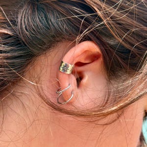 May include: A set of three silver-toned ear cuffs worn on the cartilage and lobe. The top cuff features a sunburst engraving, the middle cuff has a twisted rope texture, and the bottom cuff is a plain, smooth, thin metal band in a circular shape.