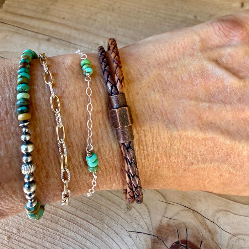 Puede incluir: Un conjunto de cuatro pulseras que incluye una con cuentas de turquesa y esferas plateadas, una cadena de eslabones ovalados dorados, una cadena fina de plata con tres cuentas de turquesa y un cord&oacute;n de cuero trenzado marr&oacute;n con cierre.