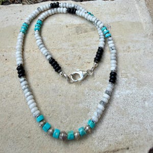 May include: A beaded necklace featuring white, black, and turquoise beads. The necklace has a silver clasp and is made of small, round beads. The design incorporates sections of black and turquoise beads, adding contrast to the white beads.