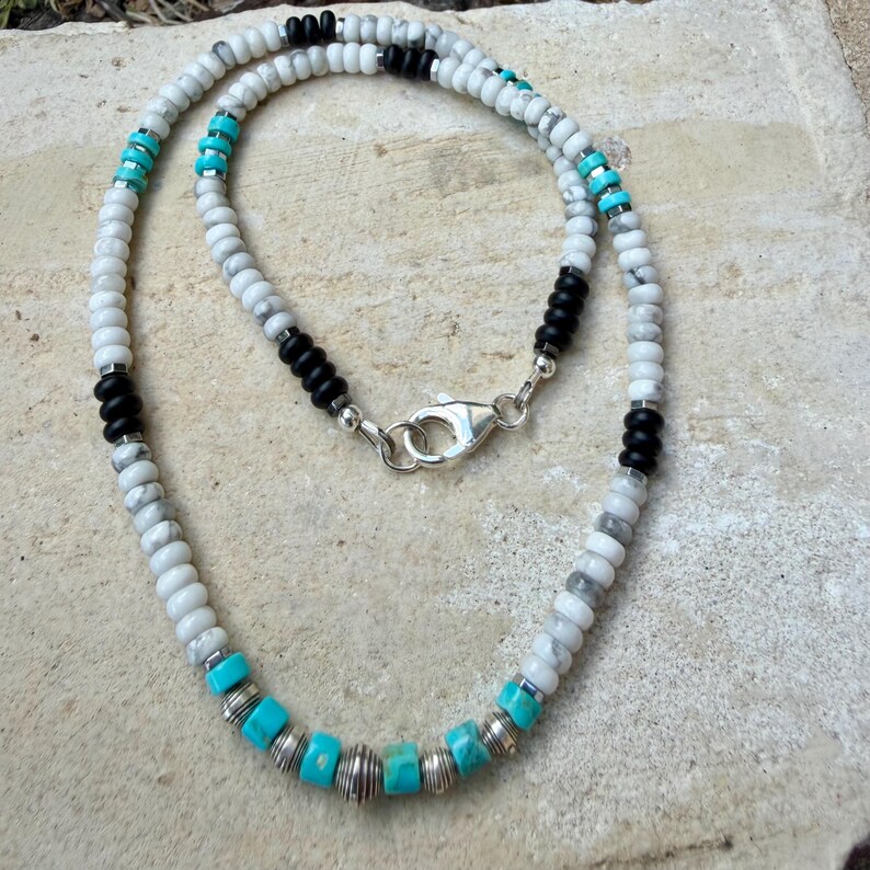 May include: A beaded necklace featuring white, black, and turquoise beads. The necklace has a silver clasp and is made of small, round beads. The design incorporates sections of black and turquoise beads, adding contrast to the white beads.