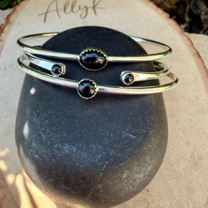 Op de afbeelding: Twee zilverkleurige armbanden met zwarte edelsteenaccenten. E&eacute;n armband heeft een grote, geschulpte zwarte steen, de andere heeft kleinere, ronde zwarte stenen. De armbanden worden getoond op een donkergrijze steen, met de naam "Allyk" op een houten oppervlak.