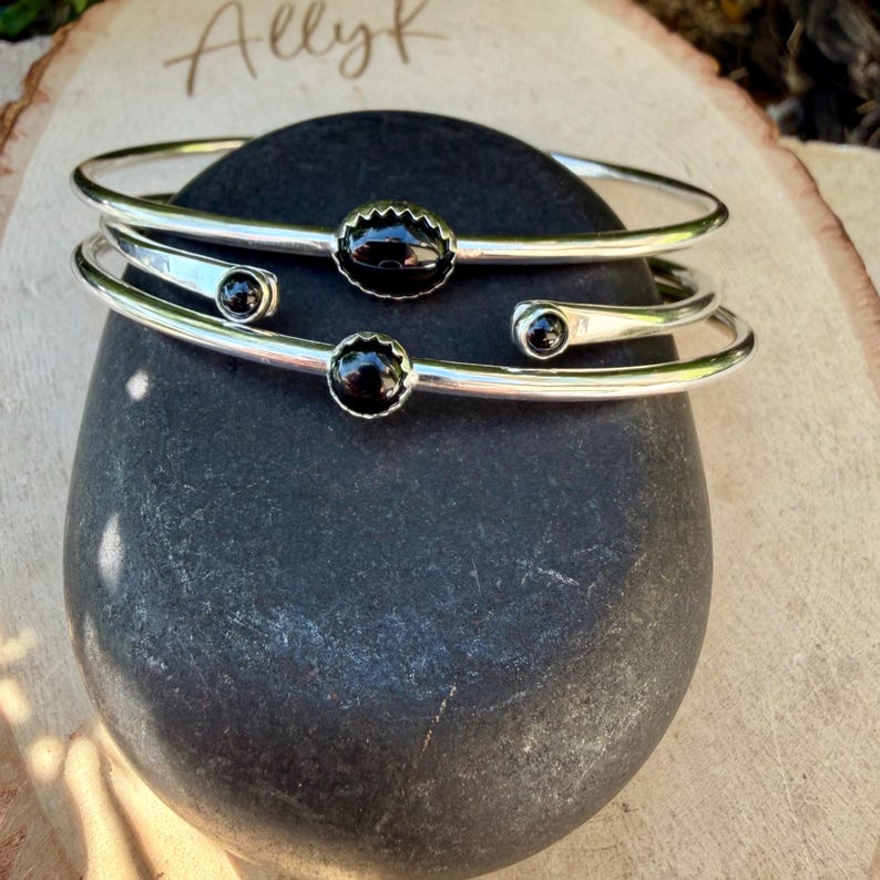Op de afbeelding: Twee zilverkleurige armbanden met zwarte edelsteenaccenten. E&eacute;n armband heeft een grote, geschulpte zwarte steen, de andere heeft kleinere, ronde zwarte stenen. De armbanden worden getoond op een donkergrijze steen, met de naam "Allyk" op een houten oppervlak.