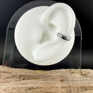 May include: A silver-toned ear cuff with an ornate, swirling design. The cuff is shown on a white ear model, set against a dark background. The jewelry piece is designed to be worn on the cartilage of the ear.
