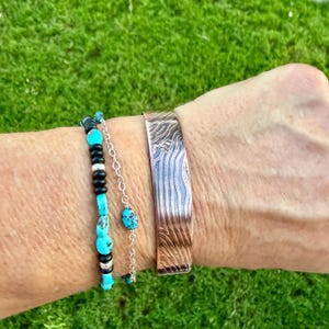 May include: A copper cuff bracelet with a textured, wave-like pattern is worn with a turquoise and black beaded bracelet. The bracelets are displayed against a green background, showcasing the jewelry's design and color.
