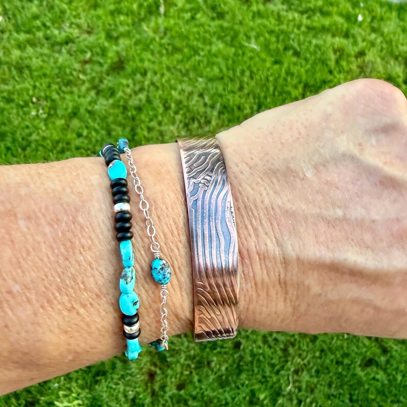 May include: A copper cuff bracelet with a textured, wave-like pattern is worn with a turquoise and black beaded bracelet. The bracelets are displayed against a green background, showcasing the jewelry's design and color.