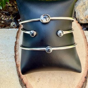 May include: Three silver-toned metal bracelets with round, speckled, black and white stones. The bracelets are displayed on a black cushion, set against a natural wood surface. The bracelets have a simple, elegant design.