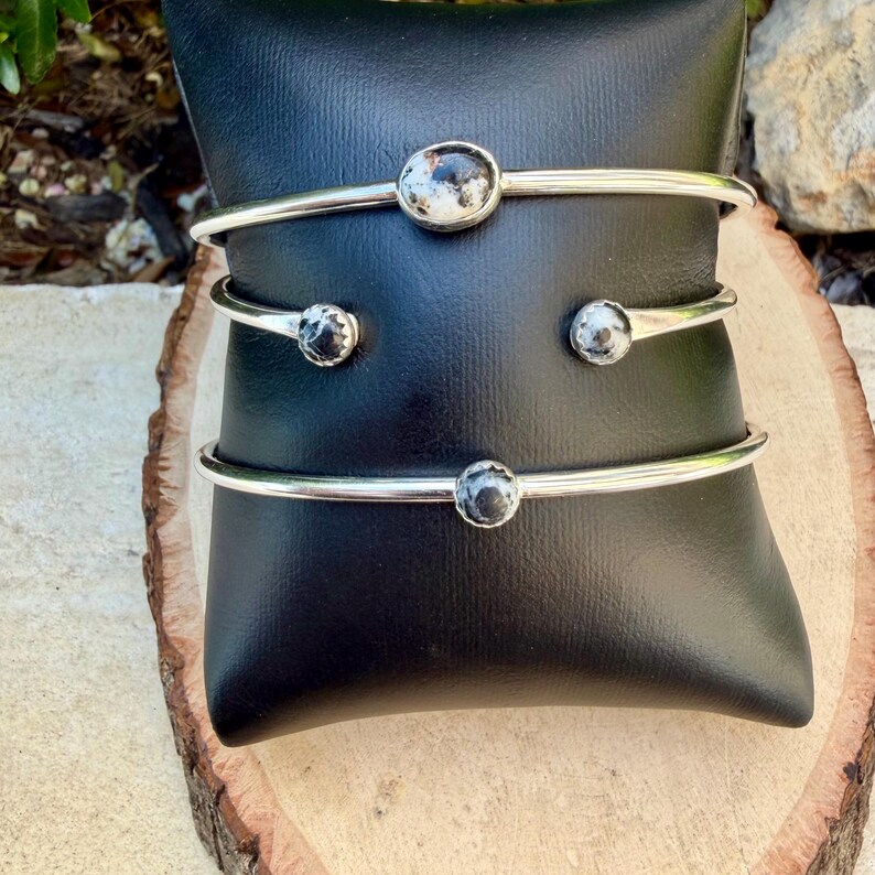 May include: Three silver-toned metal bracelets with round, speckled, black and white stones. The bracelets are displayed on a black cushion, set against a natural wood surface. The bracelets have a simple, elegant design.