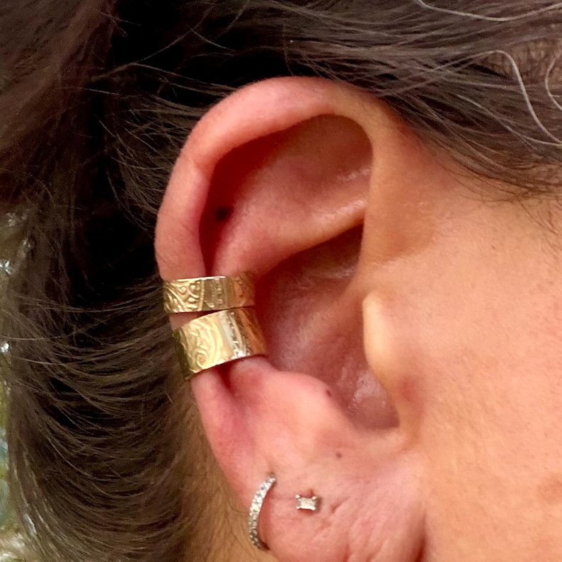 May include: Close-up of an ear adorned with two gold ear cuffs and a small, sparkling hoop earring. The ear cuffs are textured with an engraved pattern. A small, square-shaped stud earring is also visible.
