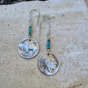 May include: A pair of silver earrings featuring vintage buffalo nickel coins. Each earring has a turquoise bead accent and a silver hook. The coins display the text "FIVE CENTS" and "UNITED STATES OF AMERICA".