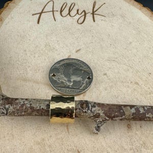 May include: A gold-coloured, hammered-texture ring sits on a small branch, next to a nickel coin. The ring has a wide band and a textured surface. The word "Allyk" is written in cursive on the wooden background.