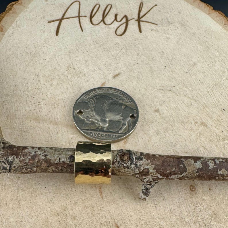 May include: A gold-coloured, hammered-texture ring sits on a small branch, next to a nickel coin. The ring has a wide band and a textured surface. The word "Allyk" is written in cursive on the wooden background.
