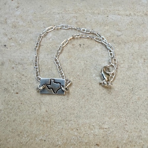 May include: A silver necklace with a rectangular pendant showcasing a cutout of the state of Texas. The pendant is linked to a chain with a lobster clasp. The necklace is presented on a neutral surface.