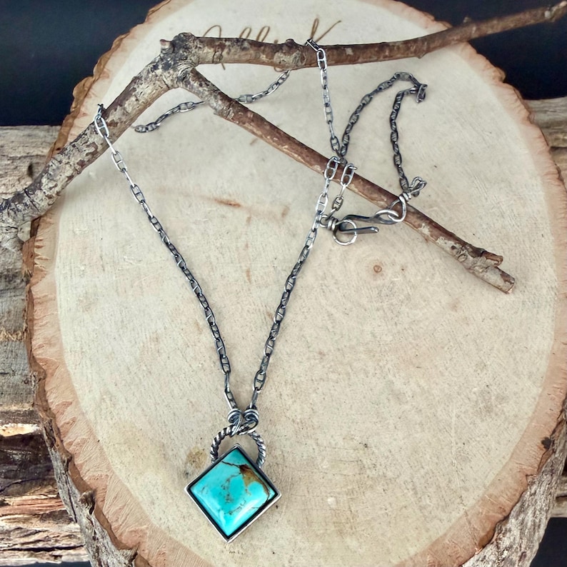 May include: A silver necklace with a turquoise stone pendant. The pendant is a square shape with a silver border and a twisted silver bail. The chain is a delicate link style with a clasp closure. The necklace is displayed on a wooden surface.