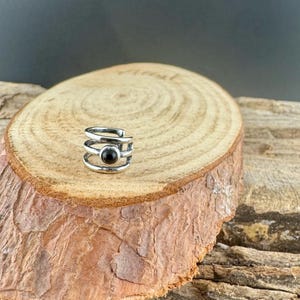 May include: A silver-colored ear cuff with a black stone set in the center. The cuff has a layered design with three bands and a curved top. The jewelry is displayed on a wooden surface with a natural, rustic appearance.