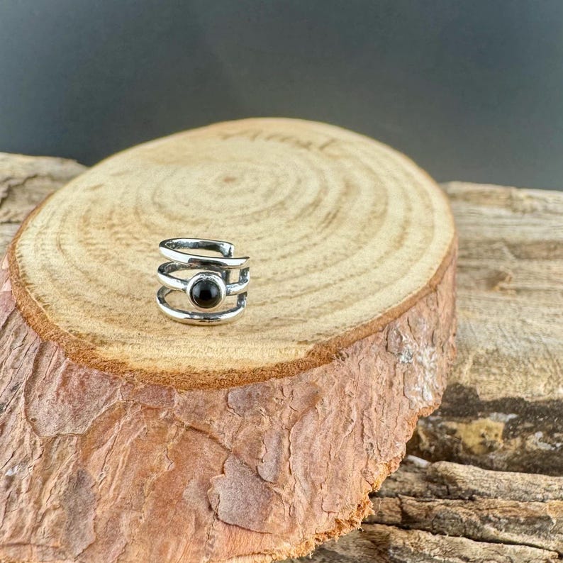 May include: A silver-colored ear cuff with a black stone set in the center. The cuff has a layered design with three bands and a curved top. The jewelry is displayed on a wooden surface with a natural, rustic appearance.