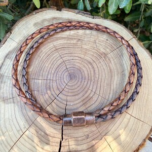 Puede incluir: Un collar de cord&oacute;n de cuero trenzado marr&oacute;n con un cierre met&aacute;lico cil&iacute;ndrico de acabado cobrizo texturizado. El cord&oacute;n est&aacute; dispuesto en forma circular, mostrando el intrincado patr&oacute;n tejido de las hebras de cuero en todo el dise&ntilde;o.