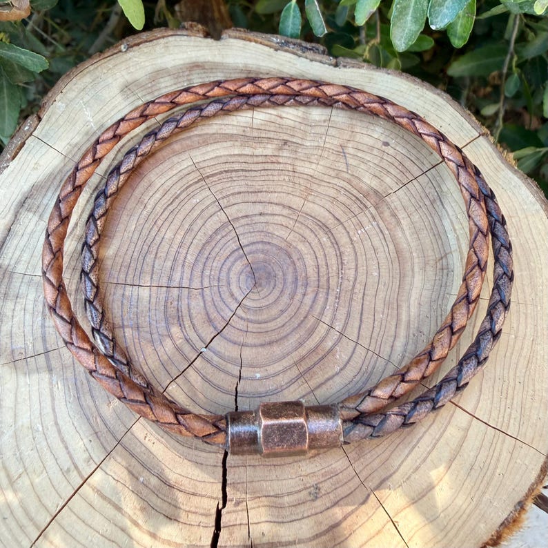 Puede incluir: Un collar de cord&oacute;n de cuero trenzado marr&oacute;n con un cierre met&aacute;lico cil&iacute;ndrico de acabado cobrizo texturizado. El cord&oacute;n est&aacute; dispuesto en forma circular, mostrando el intrincado patr&oacute;n tejido de las hebras de cuero en todo el dise&ntilde;o.