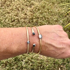 May include: Gold-toned metal bracelets with decorative black and white stone accents. The bracelets are in various styles, including a cuff and open-ended designs. The jewelry is displayed on a person's arm against a background of green grass.
