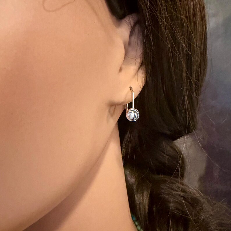 May include: Silver-tone hoop earrings with a small, round, marbled stone. The earrings are shown on a person's ear, with the stone displaying a mix of white and gray patterns. The design is simple and elegant.