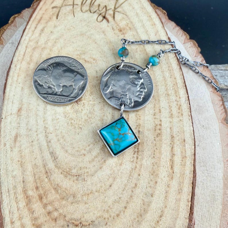 Op de afbeelding: Een zilveren ketting met een hanger van een buffel-nikkel en een turkoois edelsteen. De ketting bevat een bijpassende nikkelmunt en turkoois kralen. De sieraden worden gepresenteerd op een houten oppervlak.