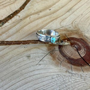 Puede incluir: Un anillo de plata con una banda pulida que presenta patrones geom&eacute;tricos grabados y una piedra turquesa de forma ovalada engastada en el centro. El anillo est&aacute; colocado alrededor de una rama de madera marr&oacute;n, mostrando el metal y la piedra.