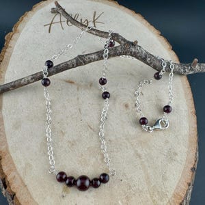 May include: A silver chain necklace with dark red garnet beads. The necklace has a delicate chain design with a series of small garnet beads interspersed along the chain. The necklace is displayed on a wooden surface with a branch.