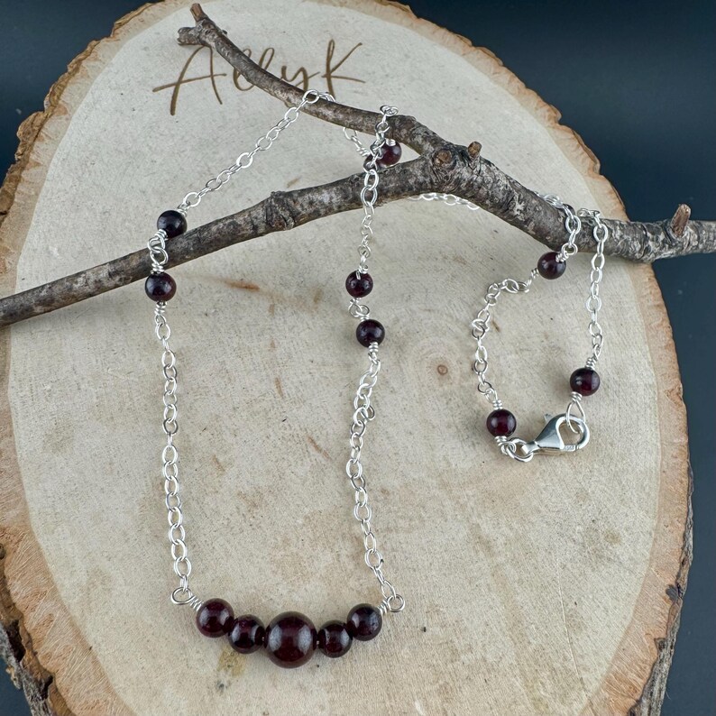 May include: A silver chain necklace with dark red garnet beads. The necklace has a delicate chain design with a series of small garnet beads interspersed along the chain. The necklace is displayed on a wooden surface with a branch.