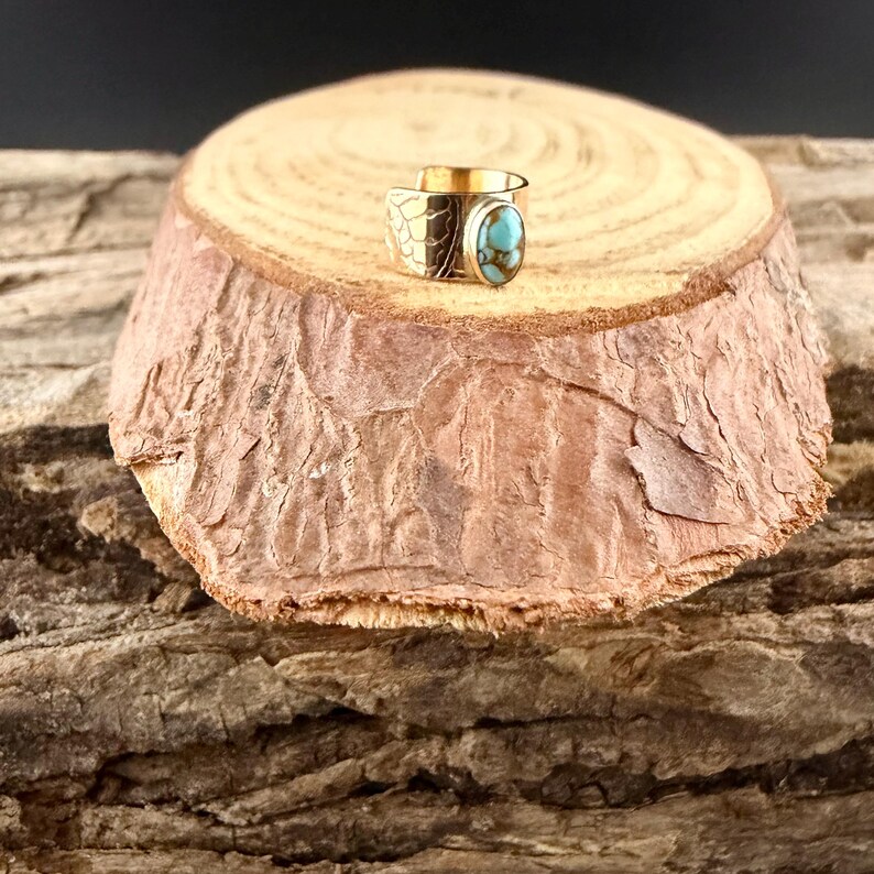 May include: A gold-toned ring with a turquoise stone and an engraved design. The ring is displayed on a wooden slice with a textured bark surface, creating a rustic aesthetic. The ring's design includes a floral pattern.