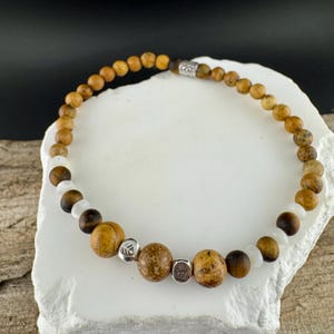 May include: A beaded bracelet featuring a mix of natural stone beads in shades of brown, tan, and white. The bracelet includes round and cylindrical beads, with small silver-colored spacers. The bracelet is displayed on a white, textured surface.
