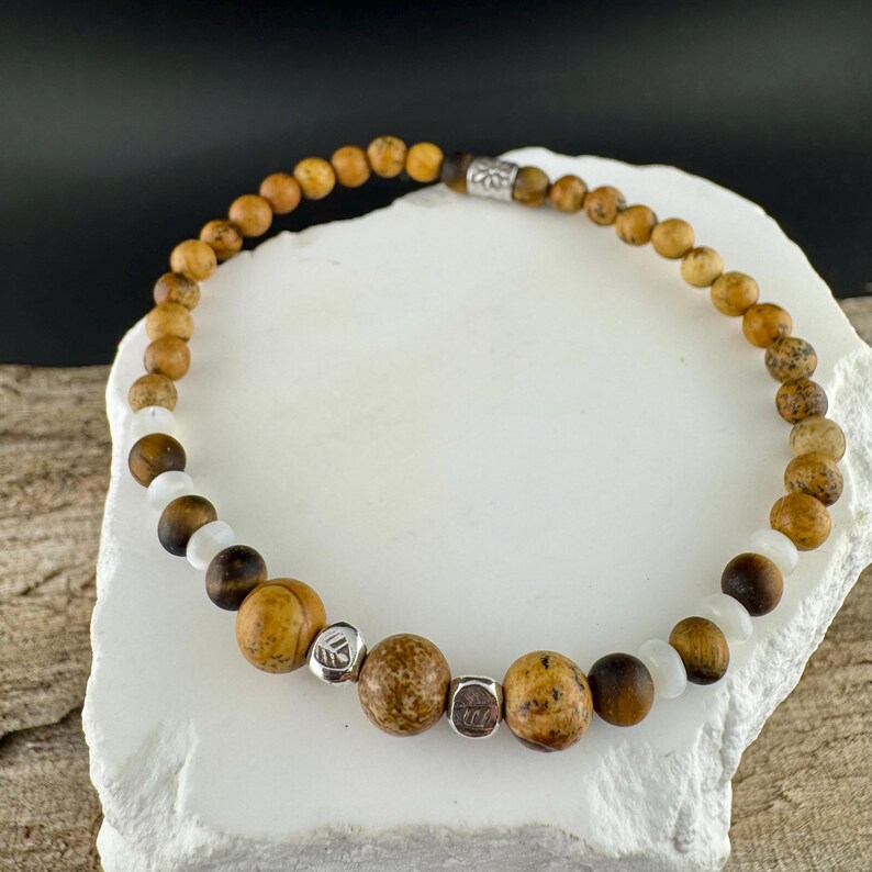 May include: A beaded bracelet featuring a mix of natural stone beads in shades of brown, tan, and white. The bracelet includes round and cylindrical beads, with small silver-colored spacers. The bracelet is displayed on a white, textured surface.