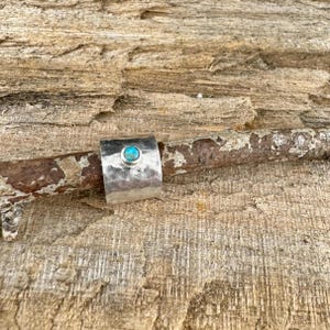 K&ouml;nnte beinhalten: Ein breiter, gehammerter Silberring mit einem t&uuml;rkisfarbenen Stein in der Mitte. Der Ring wird auf einem St&uuml;ck verwittertem Holz pr&auml;sentiert, das seine rustikale Textur und die leuchtende Farbe des Edelsteins zeigt. Ein einzigartiges Schmuckst&uuml;ck.