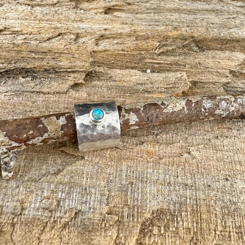 K&ouml;nnte beinhalten: Ein breiter, gehammerter Silberring mit einem t&uuml;rkisfarbenen Stein in der Mitte. Der Ring wird auf einem St&uuml;ck verwittertem Holz pr&auml;sentiert, das seine rustikale Textur und die leuchtende Farbe des Edelsteins zeigt. Ein einzigartiges Schmuckst&uuml;ck.
