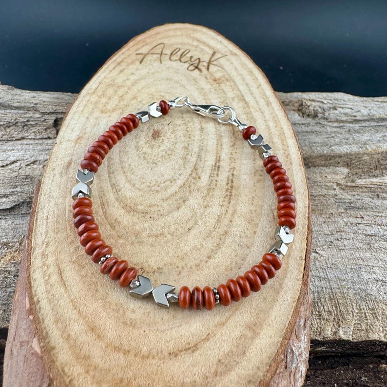 May include: A beaded bracelet featuring small, reddish-brown beads interspersed with silver-colored arrow-shaped accents. The bracelet is secured with a silver clasp. The bracelet is displayed on a wooden surface with the name "Ally K" lightly etched into the wood.