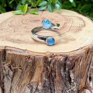 May include: A silver ring with a spiral design, featuring two round, light blue gemstones. The ring is displayed on a wooden surface, showcasing its elegant and minimalist style. The gemstones have a diameter of approximately 0.8 cm.