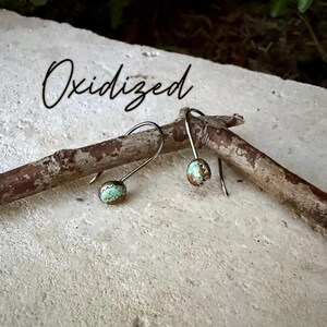 May include: A pair of drop earrings featuring small, round, light blue and brown speckled stones set in oxidized metal, hanging from curved silver-toned hooks. The word Oxidized is written in black script text above the earrings.
