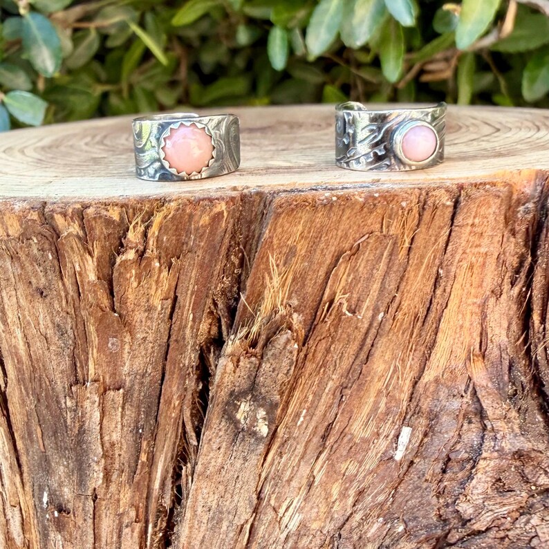 May include: Two silver rings featuring textured bands and round, light pink cabochon stones. The left ring has a scalloped bezel setting, while the right ring features a smooth, circular bezel setting. Both rings are displayed on a rough wood surface.