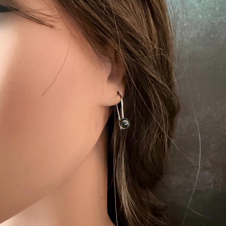 May include: A silver-toned earring featuring a long, slender U-shaped wire hook that suspends a single, small, dark, spherical bead at the base. The earring is worn in the earlobe, showcasing a minimalist design with a polished metallic finish.
