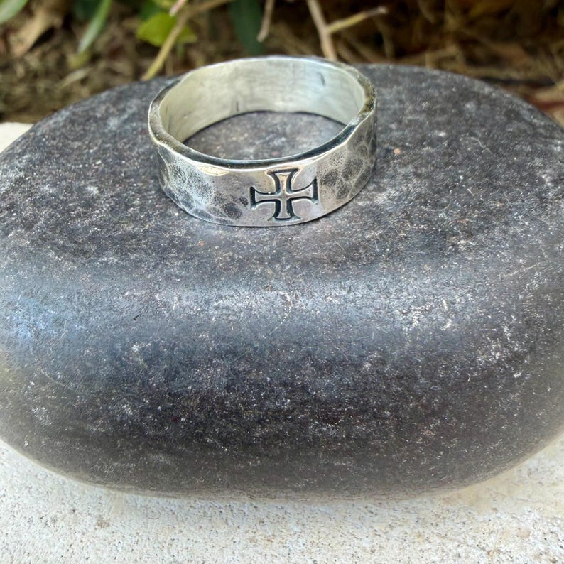 May include: A silver-toned ring with a hammered, textured finish and a prominent cross symbol engraved on the front. The band has a wide, flat profile with slightly irregular edges, showcasing a metallic sheen and a dark, recessed cross design.