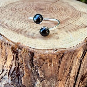May include: A silver-toned open ring featuring two round, polished black stones set at each end of the band. The ring has a bypass design with a smooth, metallic finish and two circular black cabochons that face each other with a gap in between.