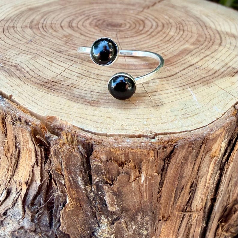 May include: A silver-toned open ring featuring two round, polished black stones set at each end of the band. The ring has a bypass design with a smooth, metallic finish and two circular black cabochons that face each other with a gap in between.