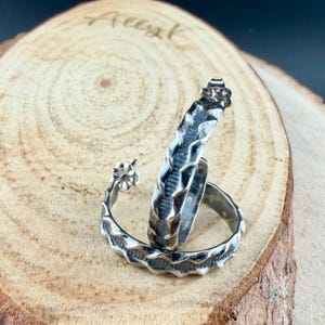 May include: A pair of silver hoop earrings with a textured, wave-like design. One earring is standing upright, showcasing the intricate pattern. The earrings are displayed on a wooden surface, highlighting their metallic sheen.