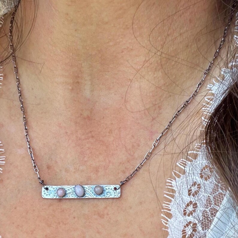 May include: A silver-toned metal necklace featuring a rectangular bar pendant with an etched scrollwork pattern. The bar is set with three small, oval-shaped, pale pink cabochon stones. The pendant hangs from a delicate, thin metal chain.
