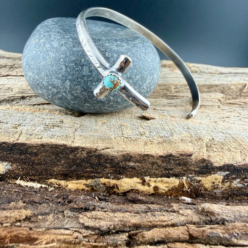 May include: A silver-toned cuff bracelet featuring a cross design with a turquoise stone center. The cross is detailed with etched patterns. The bracelet is displayed on a gray stone against a wood-grain background.