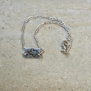 May include: A silver-toned necklace featuring a rectangular pendant with a cutout of the state of Texas. The pendant is connected to a chain-link necklace with a lobster clasp closure. The jewelry is displayed on a neutral-toned surface.